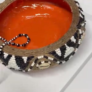 Puede incluir: Un cuenco redondo con un interior de color naranja brillante y un exterior grueso y texturizado envuelto en cuerda de yute natural. La superficie exterior está decorada con un patrón geométrico de pequeñas cuentas negras y blancas.