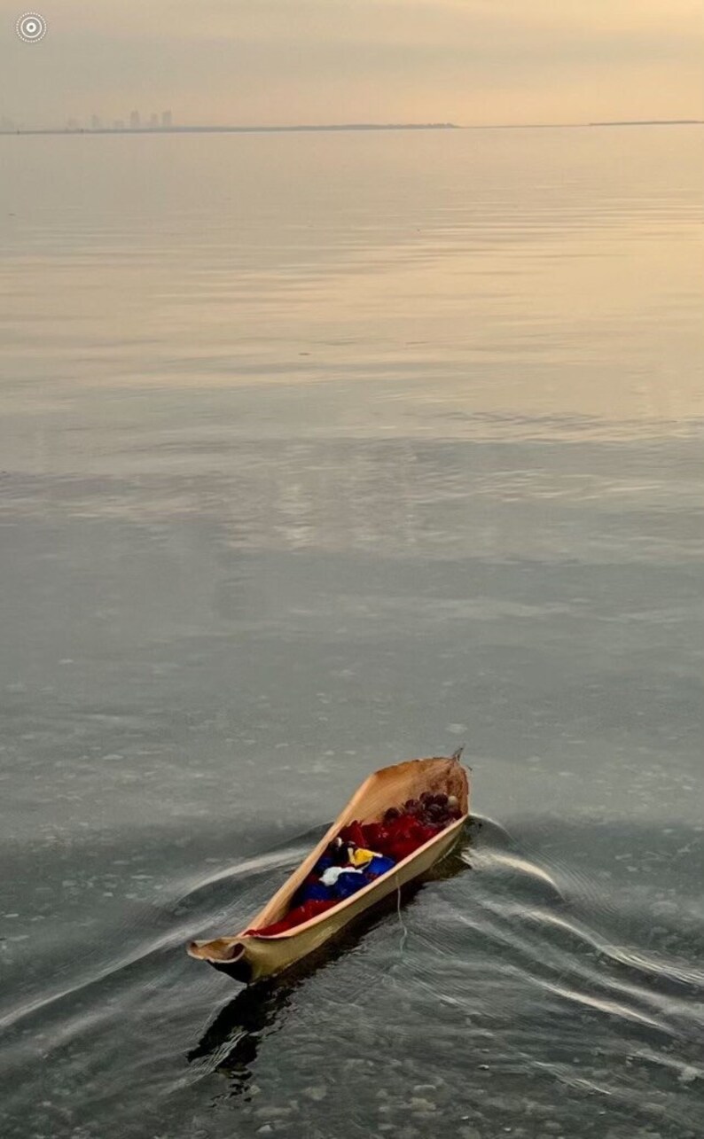 K&ouml;nnte beinhalten: Ein kleines, h&ouml;lzernes Boot schwimmt auf dem Wasser. Das Boot ist gef&uuml;llt mit roten, blauen und gelben Gegenst&auml;nden. Das Wasser ist ruhig und der Himmel ist hellblau.