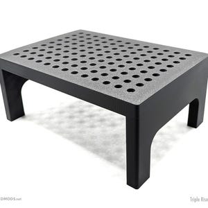 Può includere: Uno sgabello nero rettangolare con un piano grigio strutturato con numerosi piccoli fori. Lo sgabello ha quattro gambe ed è etichettato "Triple Riser - Tall". Il sito web "BOARDMODS.net" è visibile. Design moderno e funzionale.