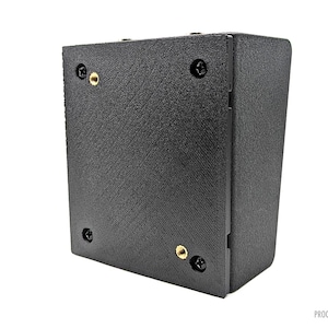 May include: A black rectangular box with a textured surface. The box has four screws and two gold-colored mounting points. The box is angled slightly to the right against a white background.