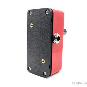 Puede incluir: Un dispositivo electrónico rectangular rojo y negro con una entrada jack plateada y un botón negro. El dispositivo tiene una superficie negra texturizada en un lado y está etiquetado como "TC-ELECTRONIC / NUX MINI".