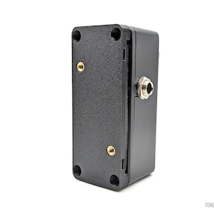 Puede incluir: Un dispositivo electrónico rectangular negro con una superficie texturizada. Tiene una entrada jack plateada y orificios para tornillos dorados. El texto "TONEX ONE" es visible.