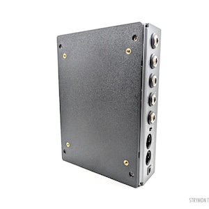 May include: A dark gray electronic device with multiple input/output jacks and a textured surface. The device has a rectangular shape with rounded corners and gold-colored screws. The text "STRYMON TRIPLE" is visible.