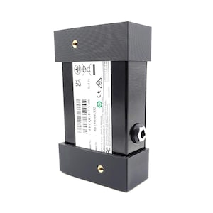 May include: A black rectangular electronic device with a label, mounted between two black rectangular brackets. The device has a small circular connector and a gold-colored screw on each bracket. The label has text and a barcode.