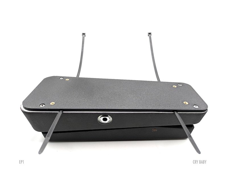 May include: Black Cry Baby guitar pedal with two black cable ties attached to the top. The pedal has a black and white stripe around the edge and a silver input jack.