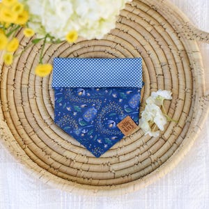 May include: A blue dog bandana with a floral pattern and a contrasting blue and white polka dot top. The bandana is displayed on a woven tray with yellow and white flowers.