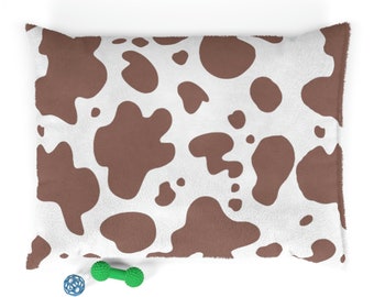May include: A rectangular pet bed with a white background and brown cow spots. A blue and green dog toy is next to the bed. The bed is made of a soft, plush material.