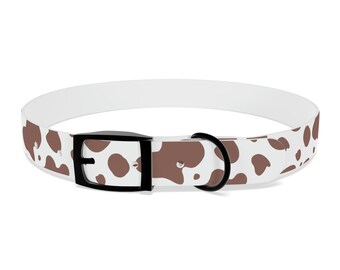 May include: A white dog collar with a brown cow print pattern. The collar has a black buckle and a black metal loop for attaching a leash. The collar is designed for pets.