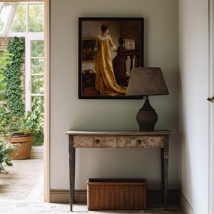 Op de afbeelding: Een gang met een ingelijst schilderij van een vrouw in een gouden jurk, een lamp met een bruine kap en een oude houten console. Een rieten mand staat eronder. De deuropening leidt naar een serre.