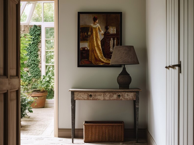 Op de afbeelding: Een gang met een ingelijst schilderij van een vrouw in een gouden jurk, een lamp met een bruine kap en een oude houten console. Een rieten mand staat eronder. De deuropening leidt naar een serre.
