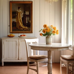 Op de afbeelding: Een lichte eetkamer met een ronde witte tafel en twee stoelen. Een vaas met oranje bloemen staat op de tafel. Een ingelijst schilderij hangt aan de muur boven een witte kast. De kamer heeft een vintage, rustieke uitstraling.