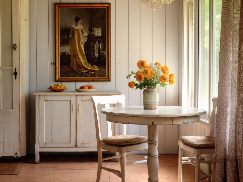 Op de afbeelding: Een lichte eetkamer met een ronde witte tafel en twee stoelen. Een vaas met oranje bloemen staat op de tafel. Een ingelijst schilderij hangt aan de muur boven een witte kast. De kamer heeft een vintage, rustieke uitstraling.