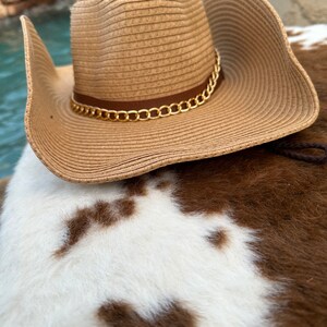 May include: A brown straw cowboy hat with a gold chain trim. The hat is sitting on a white and brown cowhide rug.