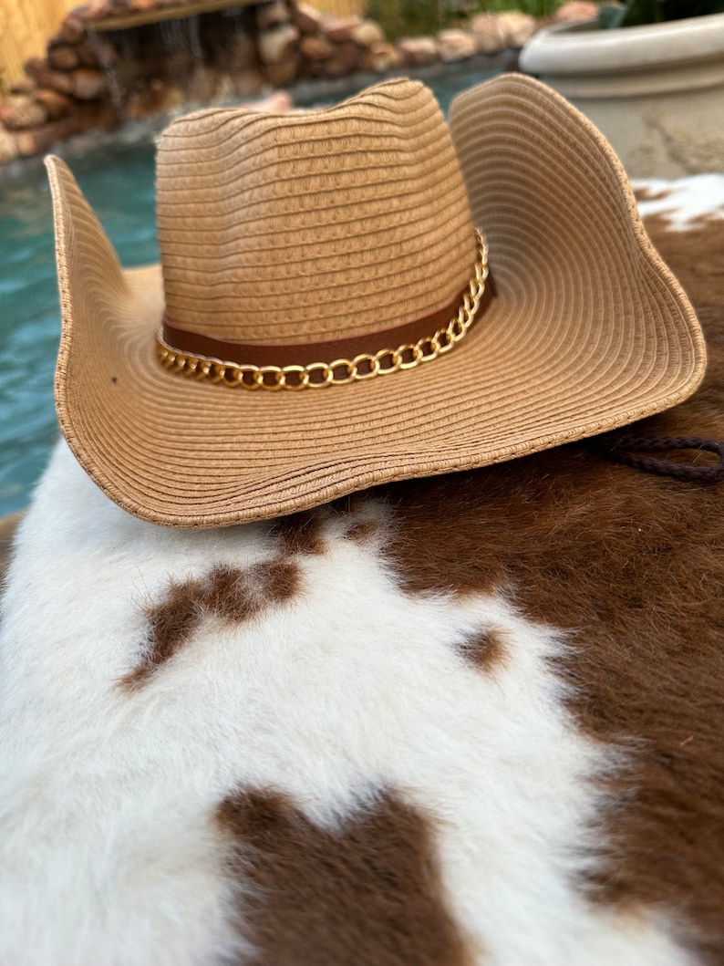 May include: A brown straw cowboy hat with a gold chain trim. The hat is sitting on a white and brown cowhide rug.