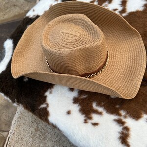 May include: A brown straw cowboy hat with a gold chain trim. The hat is sitting on a white and brown cowhide rug.