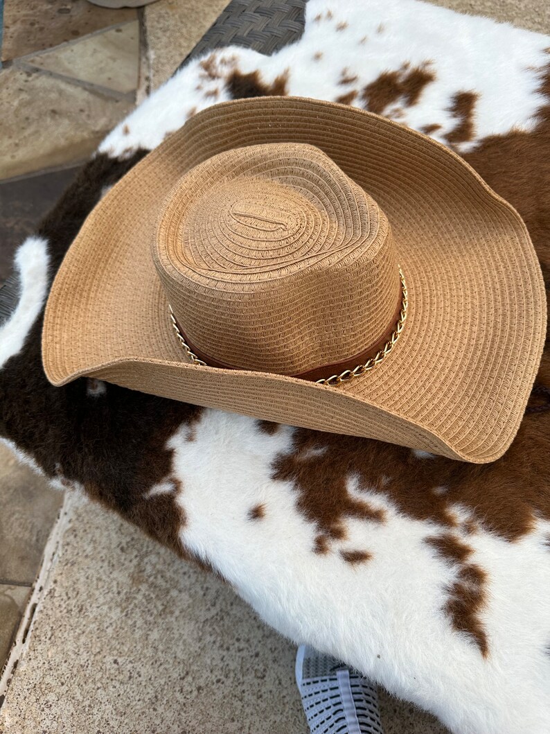 May include: A brown straw cowboy hat with a gold chain trim. The hat is sitting on a white and brown cowhide rug.