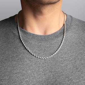 Peut inclure: Collier chaîne en argent de style corde. Le collier est porté autour du cou et repose sur un t-shirt gris. La chaîne présente un motif torsadé.
