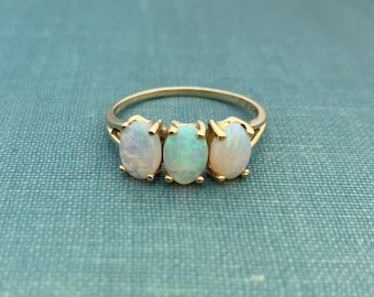 May include: A gold ring featuring three oval opals. The opals are set in a row, with a light blue-green hue in the center stone and off-white tones in the flanking stones. The ring is set against a teal background.