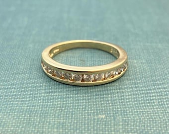 May include: A gold-colored ring with a channel setting, featuring a row of small, clear gemstones. The ring is set against a teal-colored background, showcasing its polished finish and the sparkle of the stones.