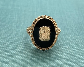 May include: A gold ring with a black oval stone set in a rope-like design. The stone features a gold crest with an eagle and shield.