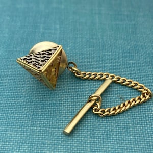 Könnte beinhalten: Ein goldfarbener Krawattenverschluss mit geometrischem Pyramidenmuster. Die Pyramide besteht aus einem gewebten Drahtgeflecht mit einer strukturierten Oberfläche. Der Krawattenverschluss ist an einer goldenen Kette mit einem Stabverschluss befestigt.