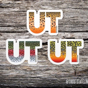 May include: Three vinyl stickers with the letters "UT" in white with a trout pattern in orange, green, and yellow. The stickers are on a wooden background. The text "@TROUTSTATELINES" is at the bottom of the image.