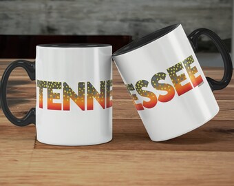 Könnte beinhalten: Zwei weiße Keramikbecher mit schwarzen Henkeln. Die Becher haben das Wort "TENNESSEE" in roten Buchstaben mit einem Forellenmuster darauf gedruckt. Die Becher stehen auf einer Holzoberfläche.