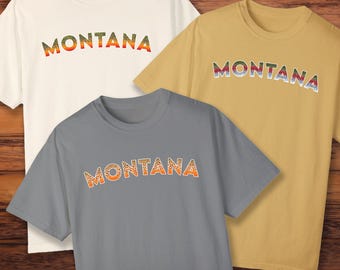 Könnte beinhalten: Drei T-Shirts mit dem Wort "MONTANA" in einem Regenbogenforellenmuster bedruckt. Die Shirts sind in Weiß, Grau und Gelb.