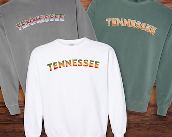 Könnte beinhalten: Drei Sweatshirts mit dem Wort "Tennessee" in einem Regenbogen-Farbverlauf auf der Vorderseite bedruckt. Die Sweatshirts sind in Grau, Weiß und Grün.