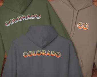 Könnte beinhalten: Drei Kapuzenpullover in Olivgrün, Grau und Beige. Die olivgrünen und grauen Sweatshirts tragen "COLORADO" auf der Vorder- und Rückseite in einer farbenfrohen, strukturierten Schrift. Das beige Sweatshirt hat "CO" auf der Vorderseite.