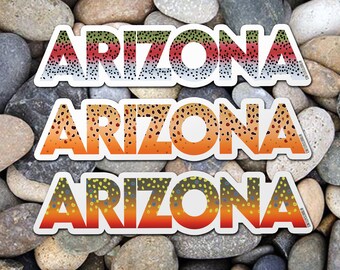 Könnte beinhalten: Drei Aufkleber mit dem Wort "Arizona" in weißen Buchstaben auf einem Hintergrund mit Forellenmuster. Der obere Aufkleber hat ein Regenbogenforellenmuster, der mittlere Aufkleber ein Braunforellenmuster und der untere Aufkleber ein Bachforellenmuster. Die Aufkleber befinden sich auf einem Hintergrund aus kleinen, glatten, grauen Steinen.