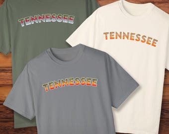 Könnte beinhalten: Drei T-Shirts mit dem Wort "Tennessee" darauf gedruckt in einem Regenbogenforellenmuster. Die Shirts sind in verschiedenen Farben: grün, weiß und grau.
