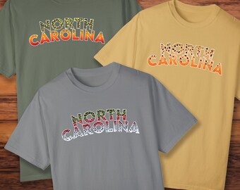 Könnte beinhalten: Drei T-Shirts mit einem Forellenmuster und dem Text "NORTH CAROLINA" in einer mehrschichtigen Schriftart. Die Shirts sind in Grün, Gelb und Grau.