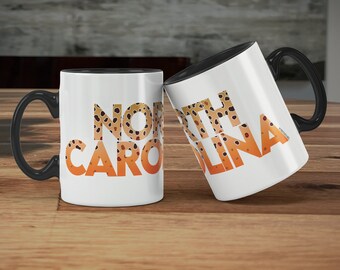 Könnte beinhalten: Zwei weiße Kaffeetassen mit schwarzen Henkeln. Die Tassen haben einen schwarzen Rand und die Worte "NORTH CAROLINA" sind in einem braunen und orangen Farbverlauf darauf gedruckt. Die Buchstaben sind mit einem Muster aus braunen Punkten gefüllt.