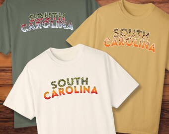 Könnte beinhalten: Drei T-Shirts mit einem South Carolina-Design. Die Shirts sind in Grün, Gelb und Weiß mit einem Forellenmuster. Der Text "South Carolina" ist in einem Regenbogen-Farbschema.