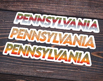 Könnte beinhalten: Drei Aufkleber mit dem Wort "PENNSYLVANIA" in einem farbenfrohen Fischschuppen-Design. Die weißen Aufkleber haben einen Farbverlauf aus Rot, Orange, Gelb und Grün. Sie liegen auf einer dunklen Holzoberfläche.