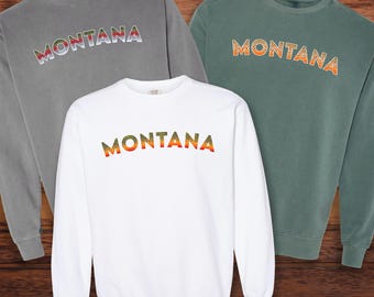 Könnte beinhalten: Drei langärmelige Sweatshirts mit dem Wort "MONTANA" in einem Regenbogen-Farbverlauf auf der Brust. Die Sweatshirts sind in Grau, Weiß und Grün.