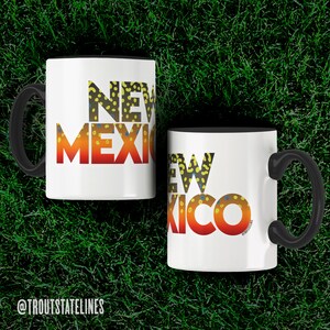 May include: Two white coffee mugs with black handles. The mugs have a black rim and the words "NEW MEXICO" printed on them in a colorful font that resembles a trout fish.