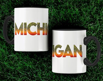 Könnte beinhalten: Zwei weiße Kaffeetassen mit schwarzen Rändern. Die Tassen haben das Wort "MICHIGAN" in einem Farbverlauf von Orange, Rot und Gelb aufgedruckt. Die Buchstaben sind mit einem Muster aus braunen und gelben Flecken gefüllt. Die Tassen stehen auf einer grünen Grasfläche. Der Text "@TROUTSTATELINES" ist unter den Tassen aufgedruckt.