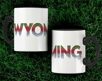 Könnte beinhalten: Zwei weiße Kaffeetassen mit schwarzen Henkeln. Die Tassen haben das Wort "WYOMING" in einem Regenbogenforellenmuster darauf gedruckt. Die Tassen stehen auf einem grünen Grasgrund.