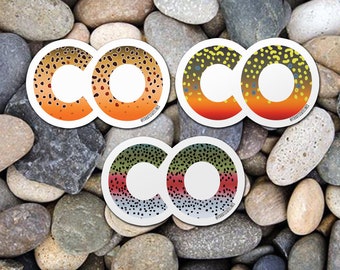 Könnte beinhalten: Zwei Sätze runder Aufkleber mit den Buchstaben "CO" in Weiß. Die Aufkleber sind mit einem Muster aus braunen, orangefarbenen, gelben, grünen und schwarzen Flecken verziert, die an die Haut einer Forelle erinnern. Der Text "@TROUTSTATELINES" ist am unteren Rand des Bildes sichtbar.