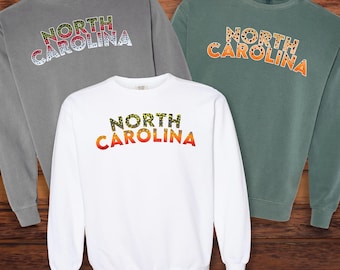 Könnte beinhalten: Drei Sweatshirts in Grau, Weiß und Grün, jedes mit "NORTH CAROLINA" in einem einzigartigen Muster. Die Sweatshirts werden auf einer Holzoberfläche präsentiert. In der unteren rechten Ecke steht der Text "@TROUTSTATELINES".