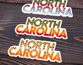 Könnte beinhalten: Drei Aufkleber mit dem Text "NORTH CAROLINA" in einem mehrschichtigen Design. Der obere Aufkleber hat ein Regenbogenforellenmuster, der mittlere ein gelb-oranges Muster und der untere ein braun-oranges Muster. Die Aufkleber befinden sich auf einer dunklen Holzoberfläche.