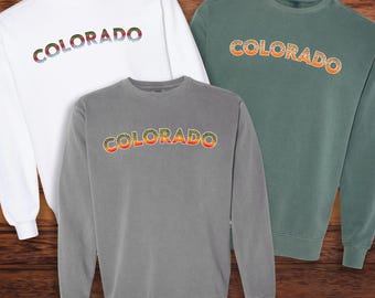 Könnte beinhalten: Drei Sweatshirts mit Rundhalsausschnitt in Weiß, Grau und Grün. Auf jedem Sweatshirt ist das Wort "COLORADO" in einem farbenfrohen Farbverlauf auf der Brust aufgedruckt. Die Sweatshirts werden auf einer Holzoberfläche präsentiert.