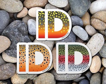 Könnte beinhalten: Drei Aufkleber mit dem Wort "ID" in weißen Buchstaben auf einem Hintergrund aus braunen, orangen und grünen Forellenmustern. Die Aufkleber sind in einer Reihe angeordnet, wobei der erste Aufkleber ein braunes Forellenmuster, der zweite Aufkleber ein oranges Forellenmuster und der dritte Aufkleber ein grünes Forellenmuster hat. Die Aufkleber befinden sich auf einem Hintergrund aus kleinen, runden, grauen Steinen.