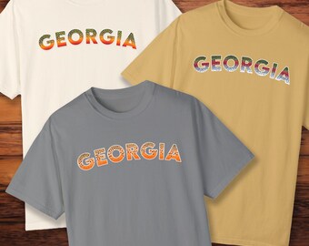 Könnte beinhalten: Drei T-Shirts mit dem Wort "GEORGIA" darauf gedruckt in einem Regenbogenforellenmuster. Die Shirts sind in Weiß, Grau und Beige.