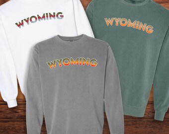 Könnte beinhalten: Drei langärmelige Sweatshirts mit dem Wort "WYOMING" in einem Regenbogenforellenmuster bedruckt. Die Sweatshirts sind in Weiß, Grau und Grün.