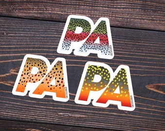 Könnte beinhalten: Drei Vinyl-Aufkleber mit dem Wort "PA PA" in einem Regenbogenforellenmuster. Die Aufkleber befinden sich auf einem braunen Holzuntergrund. Der Text "@TROUTSTATELINES" befindet sich am unteren Rand des Bildes.