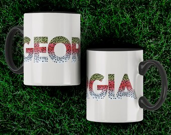 Könnte beinhalten: Zwei weiße Kaffeetassen mit schwarzen Rändern. Die Tassen haben einen schwarzen Henkel und zeigen das Wort "GEORGIA" in Regenbogenforellen-Druck.
