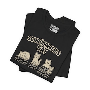 以下が含まれることがあります： ベージュ色の「Schrödinger's Cat」の文字と3匹の漫画の猫のイラストが描かれた黒いTシャツ。「Dead」「Alive」「Just Chillin'」の文字が猫の下にあります。